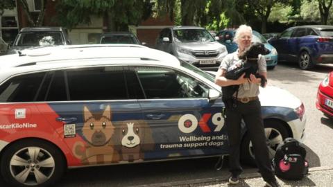 Police officer holding support dog in front of OK9 branded car.