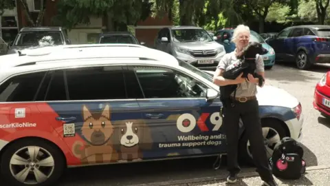 BBC Police officer holding support dog in front of OK9 branded car.