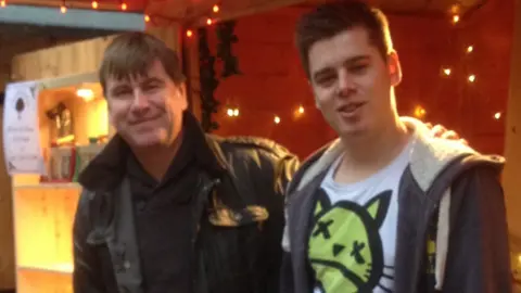 Mark Teideman Mark and his son standing in front of a market stall with white fairy lights hanging from the walls. They are smiling at the camera.