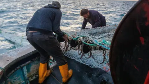 Two fishermen on a boat putting out a fishing net from their boat for their first catch of the day in the English Channel, off the south coast of East Sussex, with waves in the background and soft light from the rising sun.