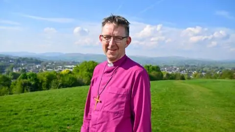 The Right Reverend Rob Saner-Haigh in a bright pink cleric's top and wearing a large crucifix. He is wearing glasses and smiling while standing on a green grassy hill above a town. There are blue skies. 