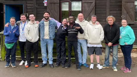 Sam Read/BBC Clients of the JH and Oarpel group are lined up with group leaders outside a brown hut posing for a photo. They are all looking at the camera, casually dressed. In the centre of them is the singer Billy Lockett. Some of them have their arms around each other. 