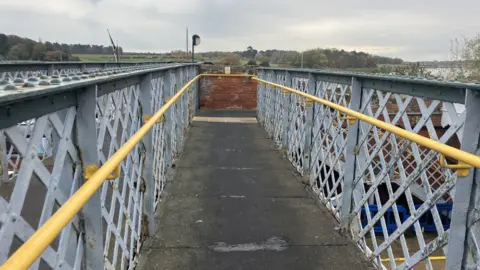 Vikki Irwin/BBC A view from on the footbridge at a railway station. The footbridge is lined with metal fences and there are yellow handrails on either side. 