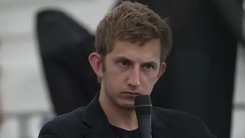 BBC Owain Williams wearing a black jacket and holding a microphone to his face