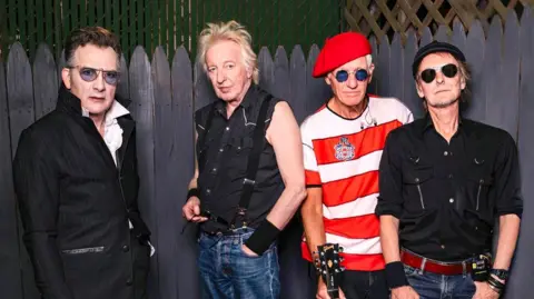 Cuffe & Taylor Four older men stand in front of a wooden fence in an outdoor space. Each has a distinct rock‑band style: one wears a long black coat with gloves and sunglasses; another wears a sleeveless black shirt with suspenders and jeans; the third wears a red beret, sunglasses, and a red‑and‑white striped shirt while holding a Gibson electric guitar; and the fourth wears a black button‑up shirt, jeans, and sunglasses.