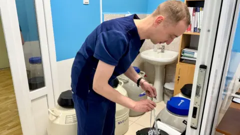 A man in blue scrubs uses a pipette at Kyiv's state-run Centre for Reproductive Medicine. There are giant white vats behind him.