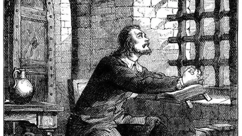 Getty Images Black and white drawing of a man with dark hair and beard looking through a grid of iron bars at a window in a prison cell. He is wearing a dark buttoned jacket with a white collar and dark trousers. His hands are resting on an open book on the window sill. There is a desk behind him with a water jar on the top. The walls are of grey brick.