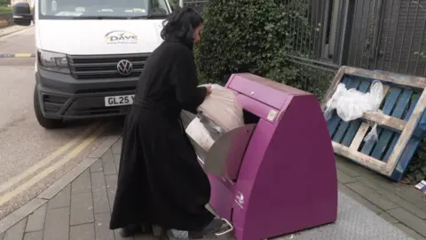 A young woman with black hair and wearing a black coat puts a bag of rubbish into a recycling chute in Tower Hamlets