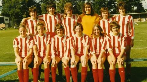 A women's football team wearing a red and white striped kit. The are stood in two rows facing the camera while on a football pitch