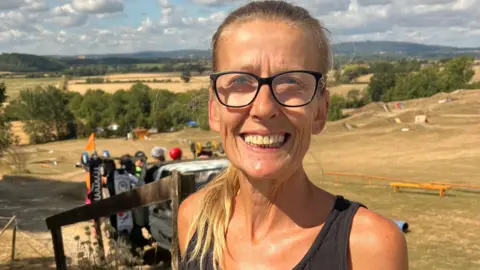 Nina smiles at the camera, from the top of the mountainboarding track which can be seen behind her. She has long blonde hair tied back in a ponytail, and wears black framed glasses and a black vest top. 