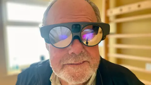 A man with a beard and wearing augmented reality glasses which have reflective lenses