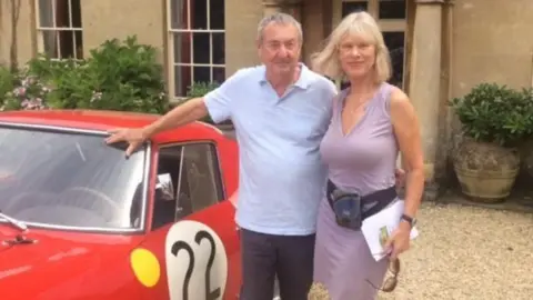 Trevor Porter A man and woman - Nick and Annette Mason - standing next to a red classic car with the number 22 on it, with sand-stone house in the background, plants and a gravel driveway.