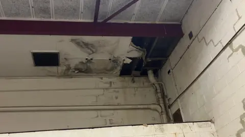 Amy Holmes/BBC An interior wall with visible damp stains and cracks, where parts of the ceiling have collapsed, exposing pipes and insulation above.