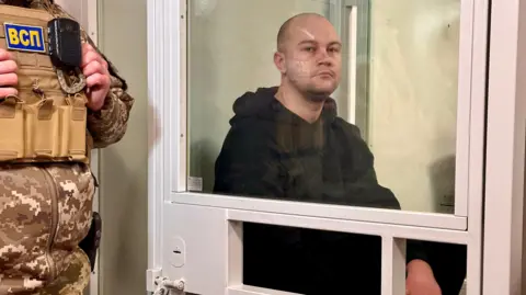 BBC/Goktay Koraltan Serhiy sits in a glass cage wearing a black hoodie, while a uniformed guard stands outside