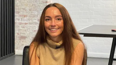 Jade has long red hair and wears a cream coloured sleeveless jumper with a high neck. She is smiling at the camera and sitting down. Behind he is a a whitewashed wall, and a ping pong table is partially visible. 