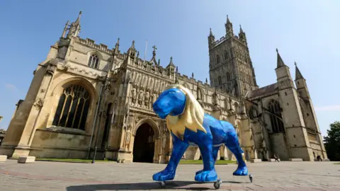 A blue lion sculpture with a gold mane outside Gloucester Cathedral on a sunny day