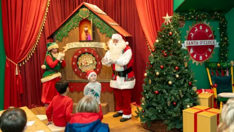 Rise Productions A grotto scene with a very large cuckoo-type clock and Santa and an elf standing by with children taking part and watching. There is a large Christmas tree to the right and a sign saying 'Santa O'Clock' in red and white