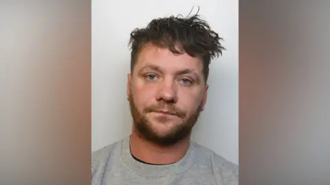 A police mugshot of Michael Ronner wearing a grey t-shirt. He has blue eyes and messy brown hair which is cut shorter on the sides. He is standing against a grey background and looking at the camera with a blank expression, his lips slightly pressed together.