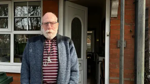 BBC Allan stands at his front door, which is slightly open showing inside his home. He wears a blue and grey cardigan, a red-and-white striped shirt, and necklaces of a cowboy boot and peace sign. He has a bald head with white beard and moustache.