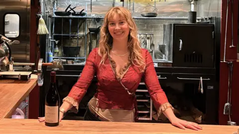 Caroline Kraetzer Caroline Kraetzer stands behind a bar, with her hands on the counter.