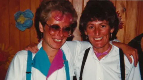 Contributed Anita and Steph in 1981, in a colour photo, Anita has on large glasses and a white top with blue and pink on, and blue braces, Steph has a white top on with black braces, they are both standing in front of a wooden panelled wall. 