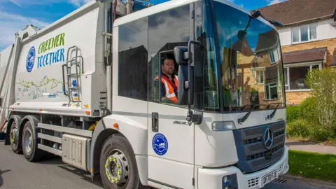 Buckingham Council A man in a large van, he is wearing an orange high-vis jacket. He is by a house. The lorry is grey in colour. 