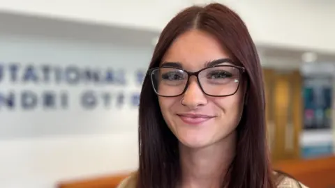 A student with dark red hair, and glasses smiling to the camera