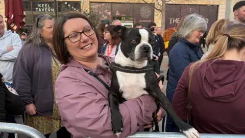 A woman in a pink coat holds a black and white Boston Terrier dog called Baxter. There are people milling around waiting for Greg James to arrive.