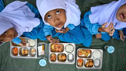 Three Indonesian school children wearing blue uniforms and white veils, looking up, as they eat a free lunch on metal trays