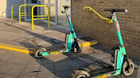 Essex County Council Two green Dott e-scooters parked outside Chelmsford railway station.