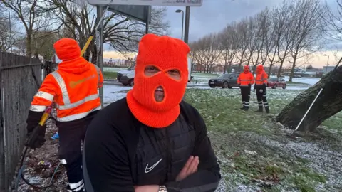 Dale Brown/Pink Mango Socials A man wearing an orange balaclava stands with his arms folded. A man behind him is cleaning a sign and another two men stand wearing orange high-vis workwear stand in the background.