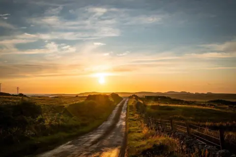 Theo Vickers A country road flanked by fields, heading into the distance, with an orange sunset in the sky.