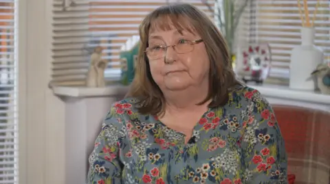 Gwen Lamb is a middle-aged woman in the grey, red and blue floral print dress. she is sitting in what appears to be a conservatory.