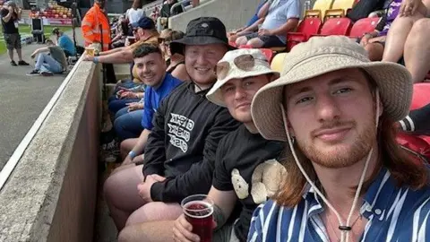 Cameron Coates The photo shows Cain North and his friends seated in a sports stadium. They are sitting in a row of red and yellow seats near the front, close to the pitch. One of them is holding a drink cup, and another is wearing a hoodie with a teddy bear design. In the background, more spectators and a steward in an orange jacket can be seen, along with part of the playing field.