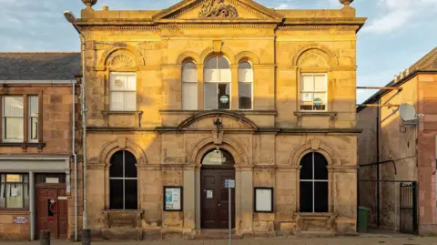 An exterior view of Invergordon Town Hall. It is a two-storey sandstone building long windows that are arch-shaped at the top. It is taller than the neighbouring properties.