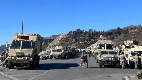 Reuters A long line of military vehicles with scattered personnel in front of the hills