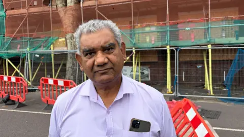 Hans Banga standing outside his destroyed building on Upper Villiers Street. He has short white hair, clean shaven and wearing a lavender shirt with the top of a smartphone poking out of a front left pocket. Behind him are railings and scaffolding.
