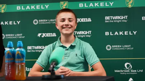 John Fairhall/BBC Ranulph, in his green polo shirt, sits smiling at the top table in the press room at Colney, where players and management conduct weekly media interviews. There is a sponsors' board behind him.