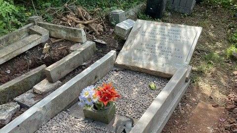 George Brunnell Rudman's headstone, with orange and purple flowers. The grave is near another broken grave and grass.