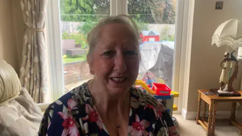 Diane is sitting on a cream coloured sofa in her living room.
The room is generally beige/cream coloured in theme.
A patio is behind her and a garden is next to the patio.