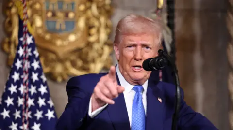 Donald Trump standing at a podium with the US flag visible behind him and a microphone in front of him - he is making a pointing gesture