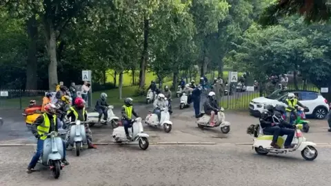 Several riders drive their scooters on the path through a park