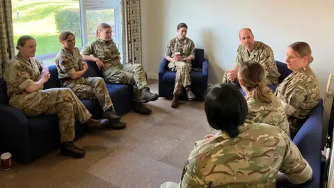Prince William, in his camouflage gear, sits on blue chair and speaks to women in military attire.