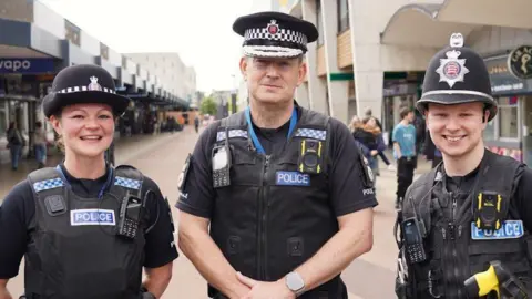 Chief Constable Ben-Julian Harrington with fellow police officers either side of him. All are wearing black police uniform and hats while standing in Basildon town centre, which is lined with shops.