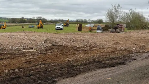 A field, which has been partially turned over to soil, has a series of yellow diggers on. To one side large brown pipes are stacked up alongside wooden beams. Pylon wires run overhead.