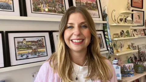 BBC Francesca Young, with shoulder-length blonde hair, is wearing a white top with pink sleeve as she smiles at the camera