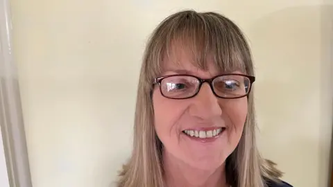 John Devine/BBC Jill Bolton, 63, has sandy shoulder length hair and has glasses with dark frames. A magnolia coloured wall is behind her.
