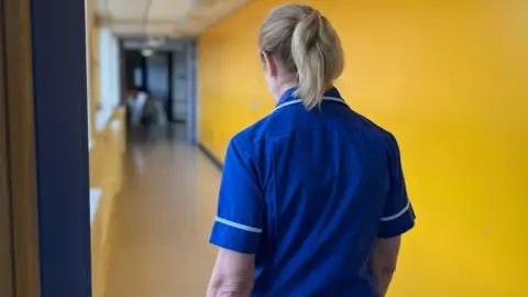 A nurse walks down a corridor in a hospital. The nurse has blonde hair tied in a ponytail. She is wearing blue scrubs. The walls in the corridor are yellow.