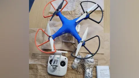 West Midlands Police A blue drone with orange and black rotor shields and white blades on a wooden table with two tinfoil wraps and a string