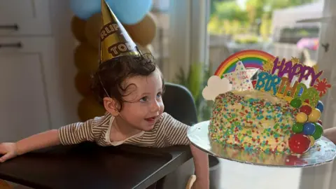 David Smith A one-year-old boy, sitting in a high chair, looking at a birthday cake. He is wearing a party hat and brown-and-white striped t-shirt. The cake is covered in colourful icing and birthday decorations.